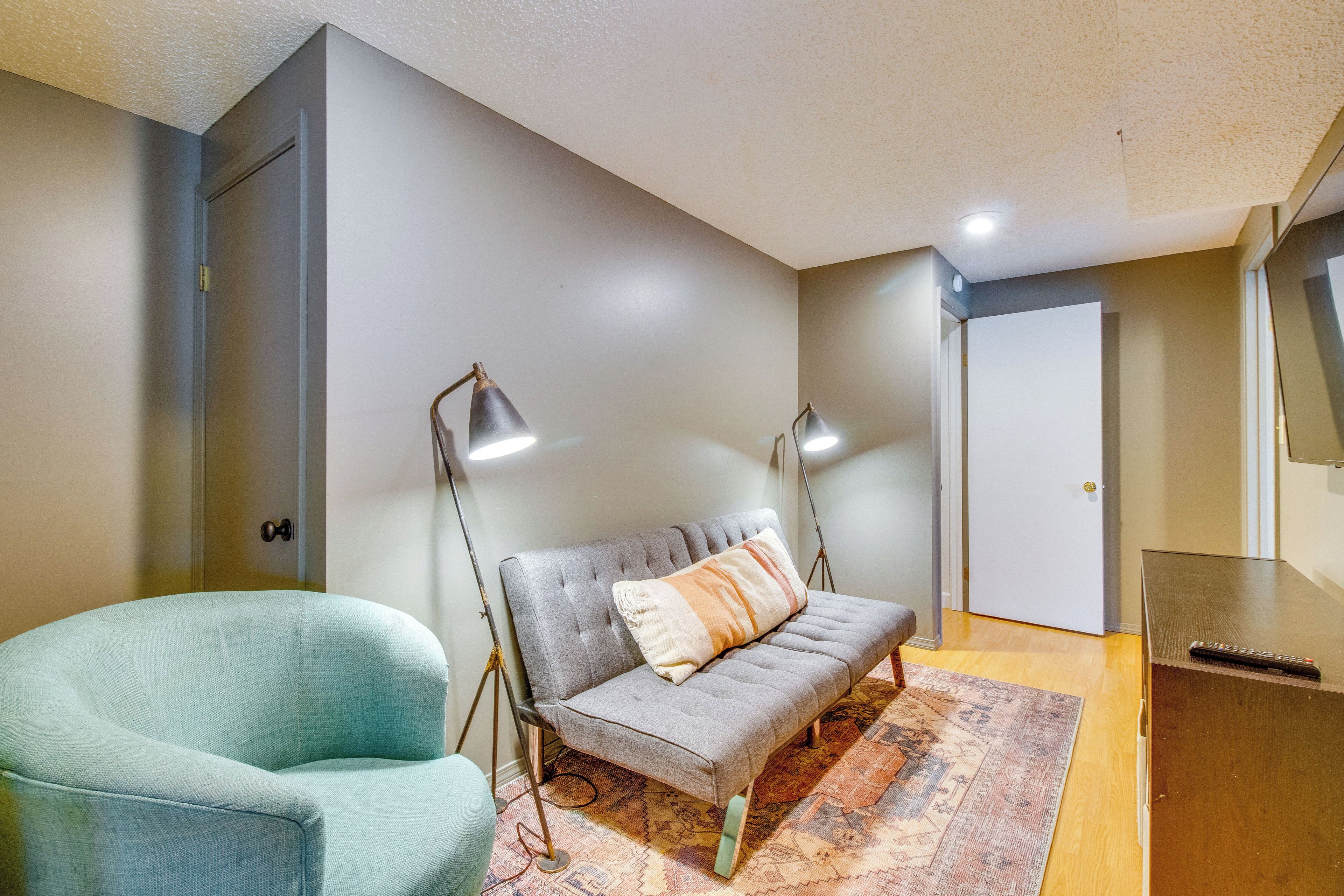 Una habitación con un sofá gris, un sillón azul verdoso y un escritorio de madera con un control remoto.