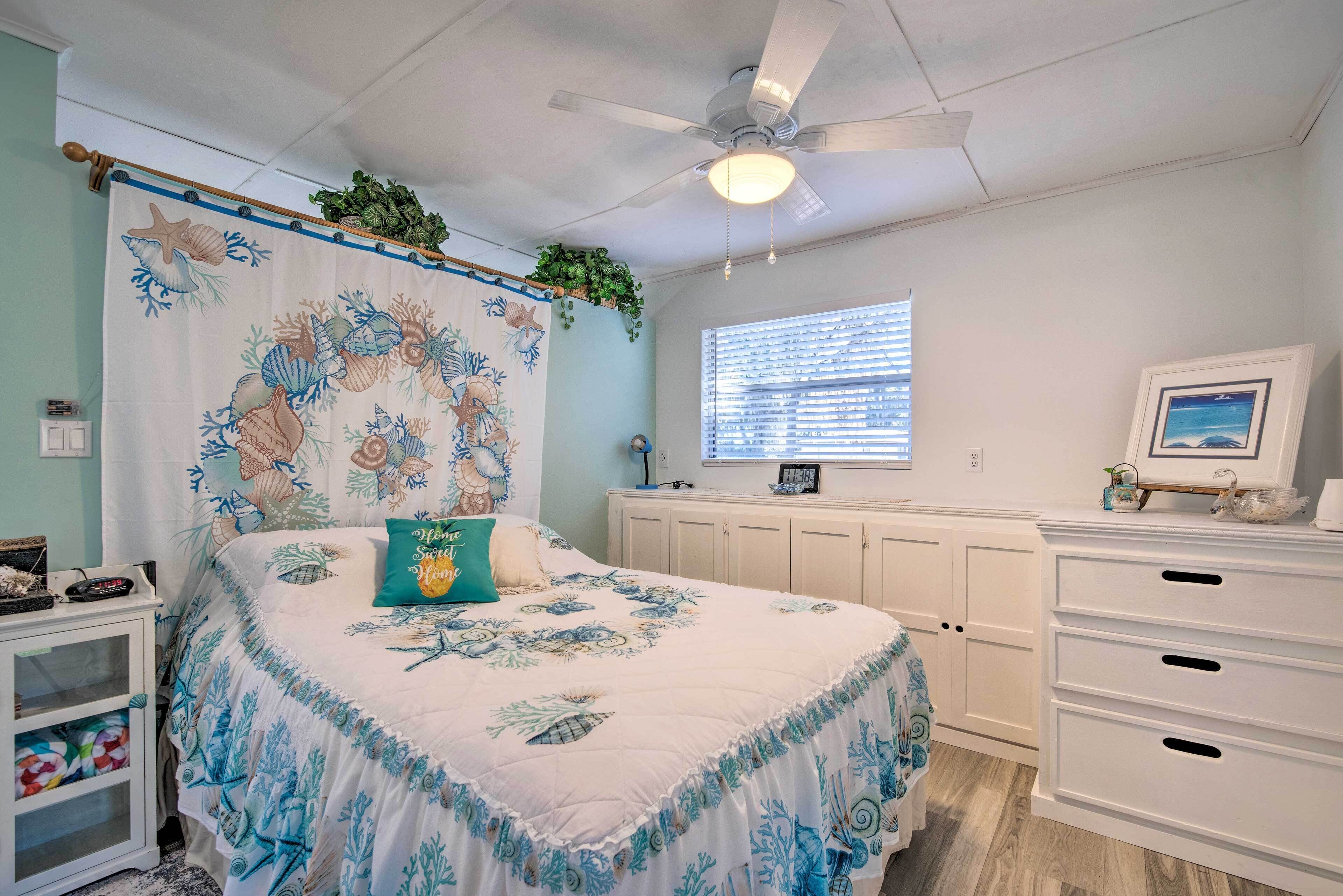 Une chambre à coucher avec un lit, un ventilateur de plafond, une fenêtre avec des stores, une commode et une plante suspendue au plafond.