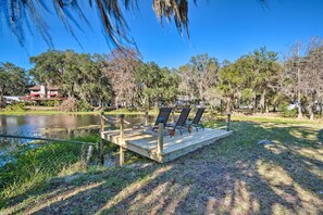 A wooden deck with chairs overlooking a pond, surrounded by trees and a building in the background. - Waterfront Studio w/ Dock Access in Inglis! (Inglis)