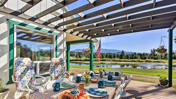 A covered outdoor dining area with a table set for a meal, chairs, and a view of a lake and mountains. - Family Home w/ Stunning River & Mountain View (Priest River)