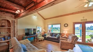 A cozy living room with wooden beams, a television, comfortable seating, and a view of the outdoors through large windows. - Family Home w/ Stunning River & Mountain View (Priest River)