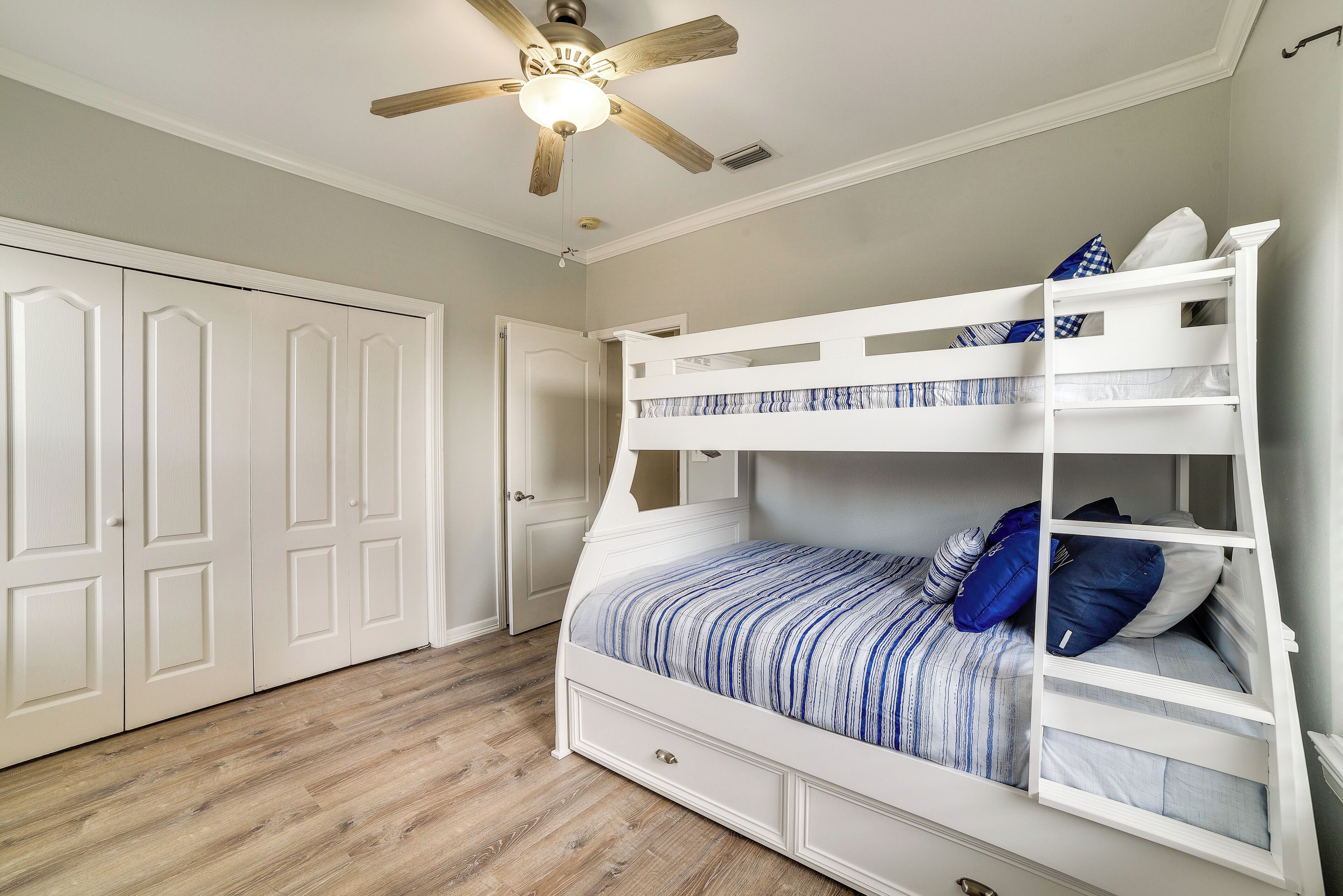 Un lit superposé avec une structure blanche, un ventilateur de plafond et un plancher en bois.