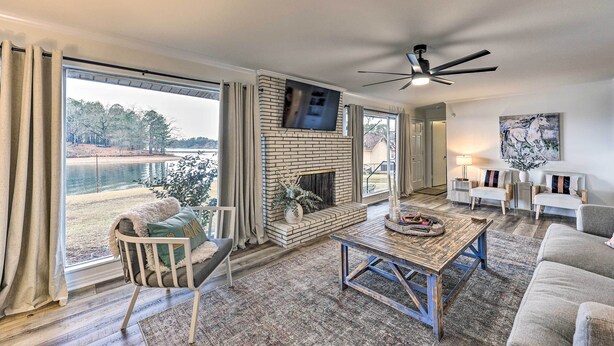 A living room with a fireplace, a large window with curtains, a ceiling fan, and a view of a body of water. - Waterfront Home w/ Dock > 3 Mi to Oaklawn Racing! (Hot Springs)