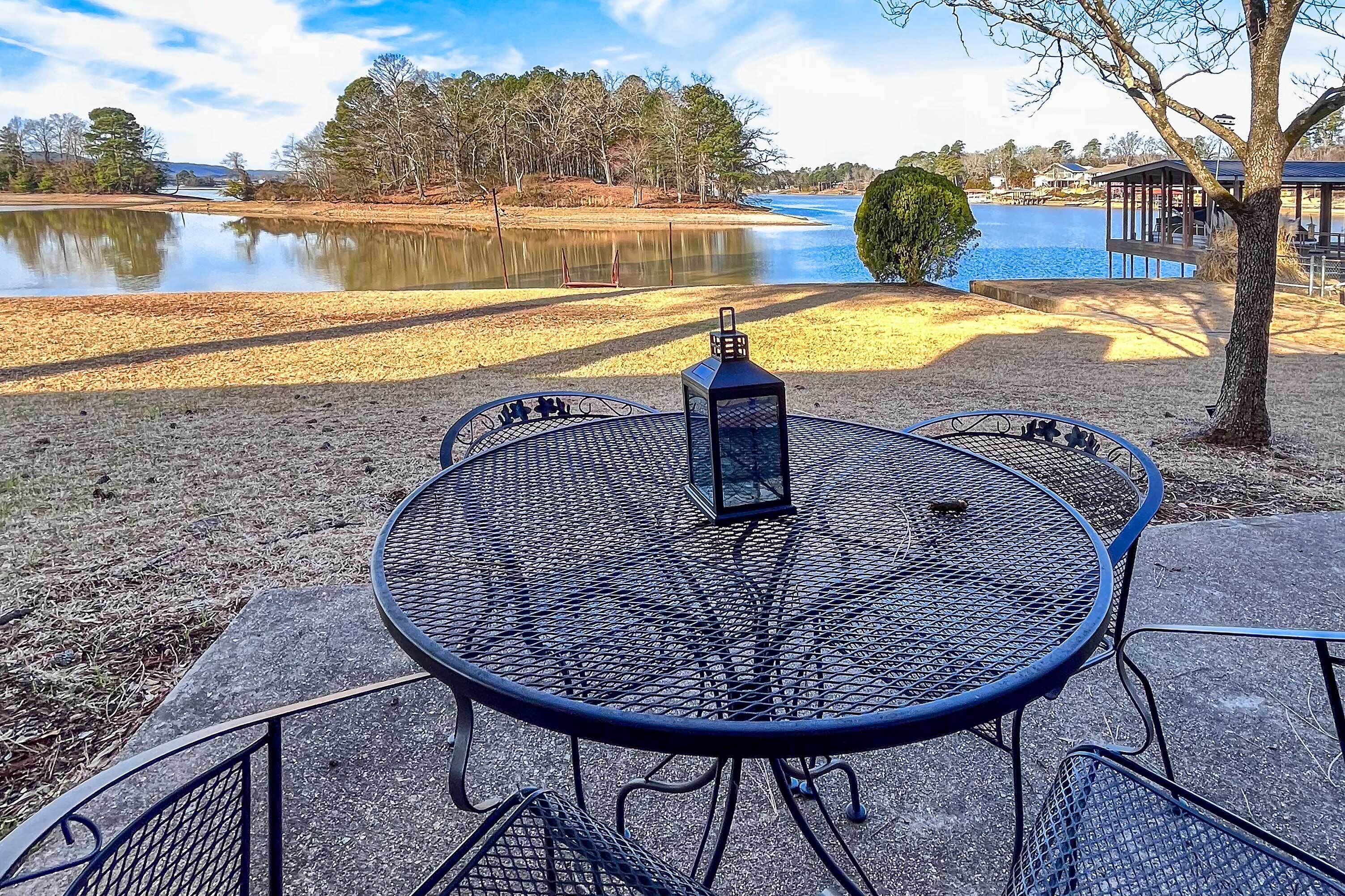 A round metal table with four chairs and a lantern, set on a concrete patio with a view of a lake and trees.