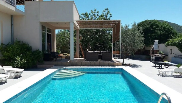 Una piscina con un pergolato e area salotto esterna, immersa nel verde e affiancata da un edificio moderno.