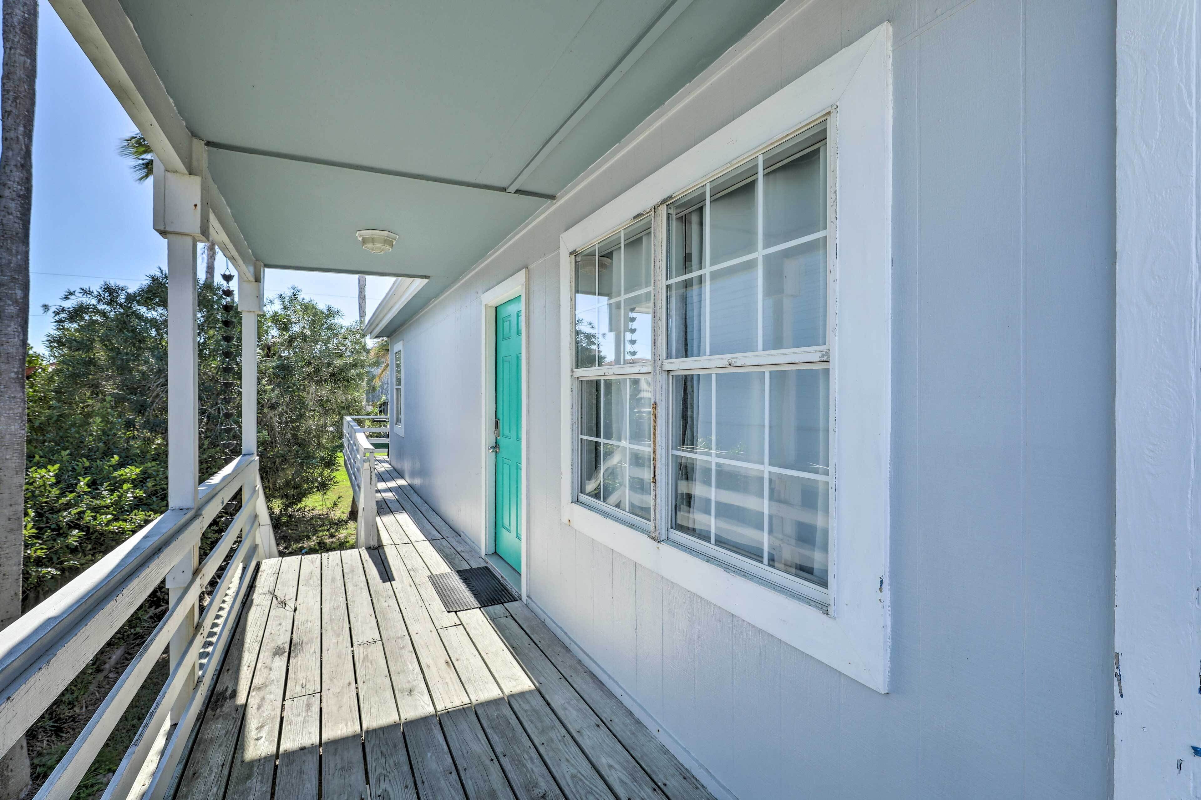 Une terrasse en bois couverte, dotée d’une balustrade blanche, d’une porte verte et d’une fenêtre aux encadrements blancs.