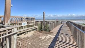 En träpromenad som leder ner till stranden, med ett träräcke på ena sidan och en informationsskylt på den andra.