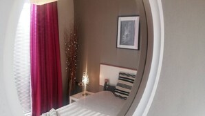 Een slaapkamer met een ronde spiegel die een bed, een nachtkastje, een lamp en een schilderij aan de muur weerspiegelt.
