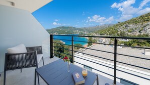 A balcony with a view of a coastal landscape, a table with a book and a glass of orange juice, and a chair with a pillow.