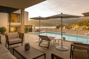 Zona junto a la piscina con sillones de descanso, sombrillas y vistas a un lago y montañas.