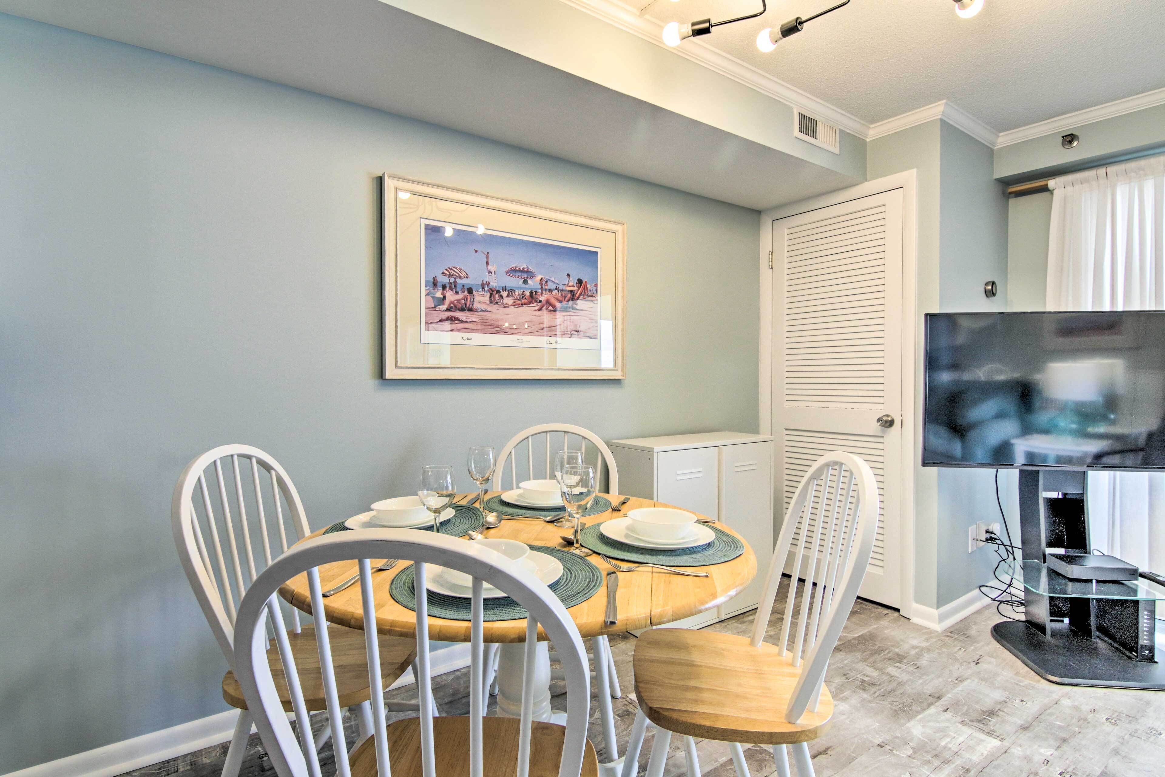 A dining area with a round table set for four, white chairs, a framed picture on the wall, and a television on a stand.
