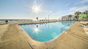 A swimming pool with clear blue water, surrounded by lounge chairs and umbrellas, with a view of the ocean and a building in the background. - Beachfront PCB Escape w/ Free Beach Chair Services (Panama City Beach)