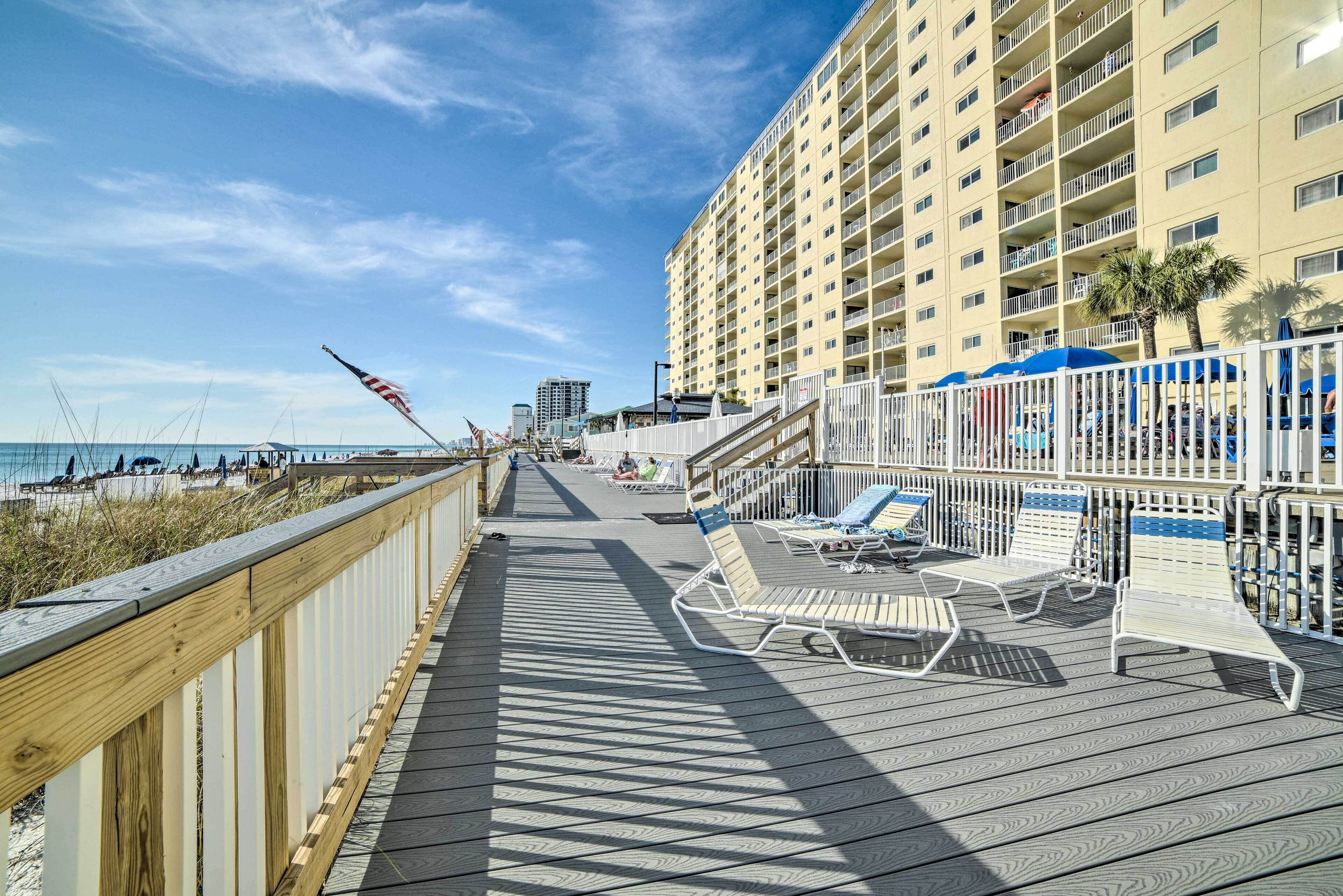 A wooden boardwalk leading to a beach with lounge chairs and umbrellas, overlooking the ocean and a high-rise building.