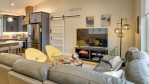 A modern living room with a leather sofa, a dining area with a table and chairs, a kitchen with grey cabinets and stainless steel appliances, and a large flat-screen TV.