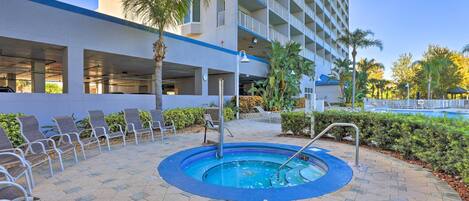 Uma área de piscina de hotel com espreguiçadeiras, uma banheira de hidromassagem e um edifício com varandas ao fundo.