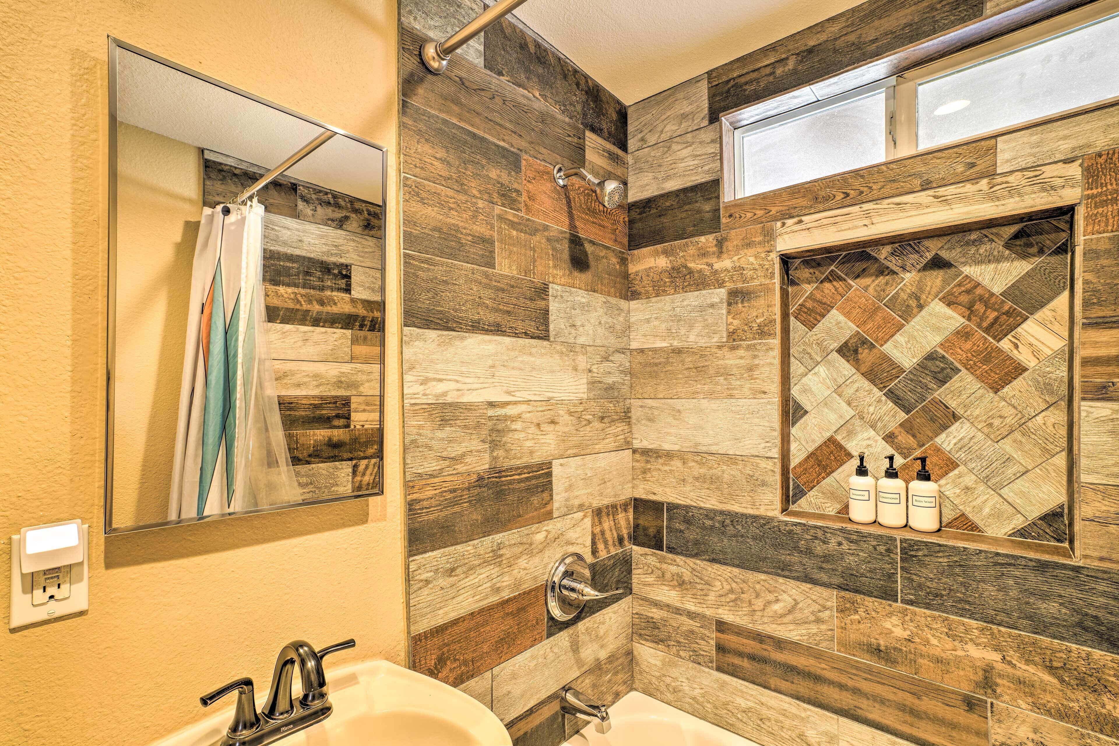 Une salle de bain avec un mur décoré d’une mosaïque en bois, un miroir, une douche, un lavabo et deux bouteilles de shampoing.