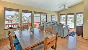 A spacious living area with a wooden dining table, grey sofa, and large windows offering a view of a red-roofed building and trees. - Pagosa Springs Vacation Rental w/ Boat Dock! (Pagosa Springs)