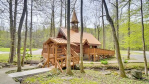 Une cabane en bois avec un toit en pente et un clocher, entourée d’arbres et d’une zone herbeuse.