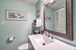 A bathroom with a white sink, a mirror, and a toilet. There is a framed picture on the wall and a basket with towels.