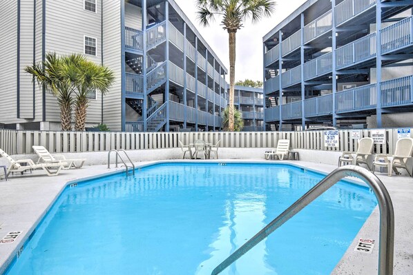 A swimming pool with lounge chairs and a lifeguard sign, surrounded by multi-story buildings.