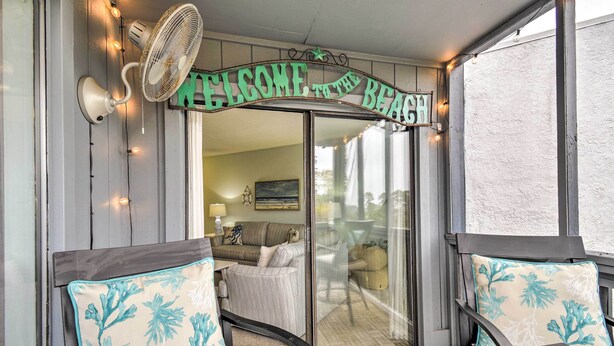 Un espace extérieur accueillant, avec des sièges confortables, une pancarte indiquant « Bienvenue à la plage », un ventilateur et une porte vitrée donnant sur un balcon.