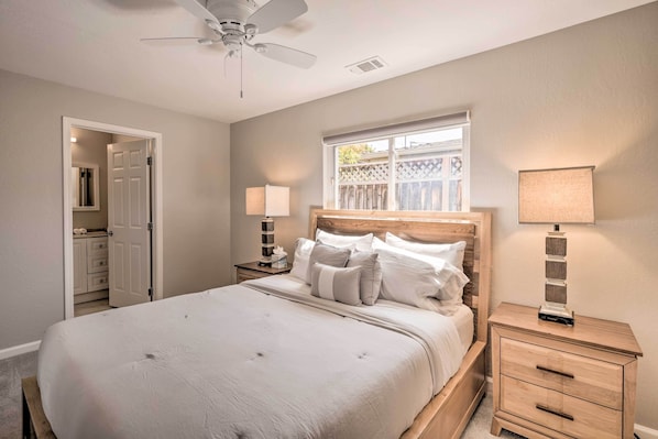 A bedroom with a large bed, a wooden headboard, two bedside tables with lamps, a ceiling fan, and a window with a view of a wooden fence.