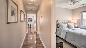A hallway with wooden flooring leading to a bedroom with a bed, a ceiling fan, and a nightstand with a lamp.