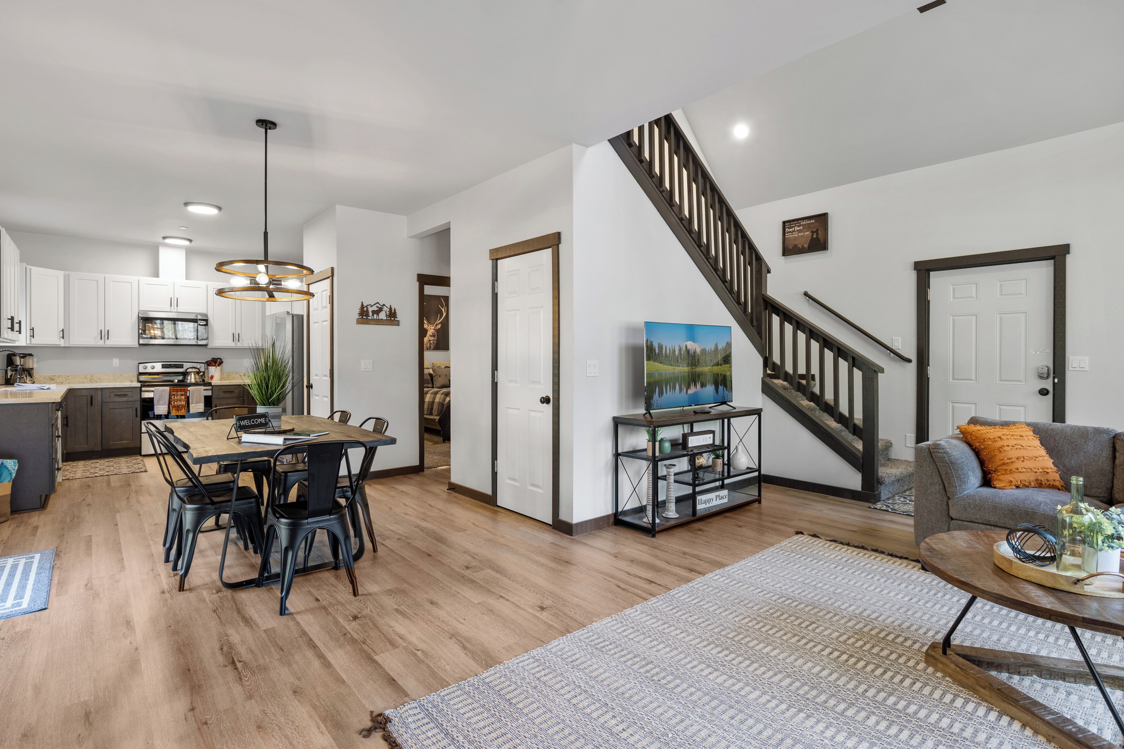 A modern kitchen with white cabinets, a dining area with a wooden table and chairs, a living room with a sofa and a TV, and a staircase with a wooden railing.