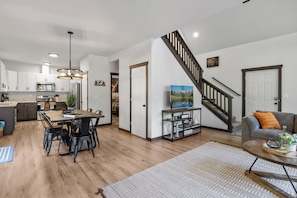 Una cocina moderna con armarios blancos, un comedor con mesa y sillas de madera, una sala de estar con sofá y televisor, y una escalera con barandilla de madera.