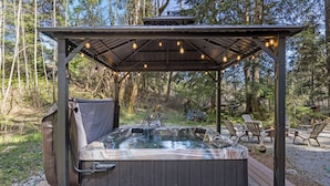 Un jacuzzi bajo una pérgola de madera con luces colgantes, rodeado por una terraza y mobiliario exterior.