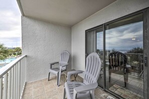 Een balkon met twee Adirondack stoelen, een glazen deur en uitzicht op een zwembad en palmbomen.
