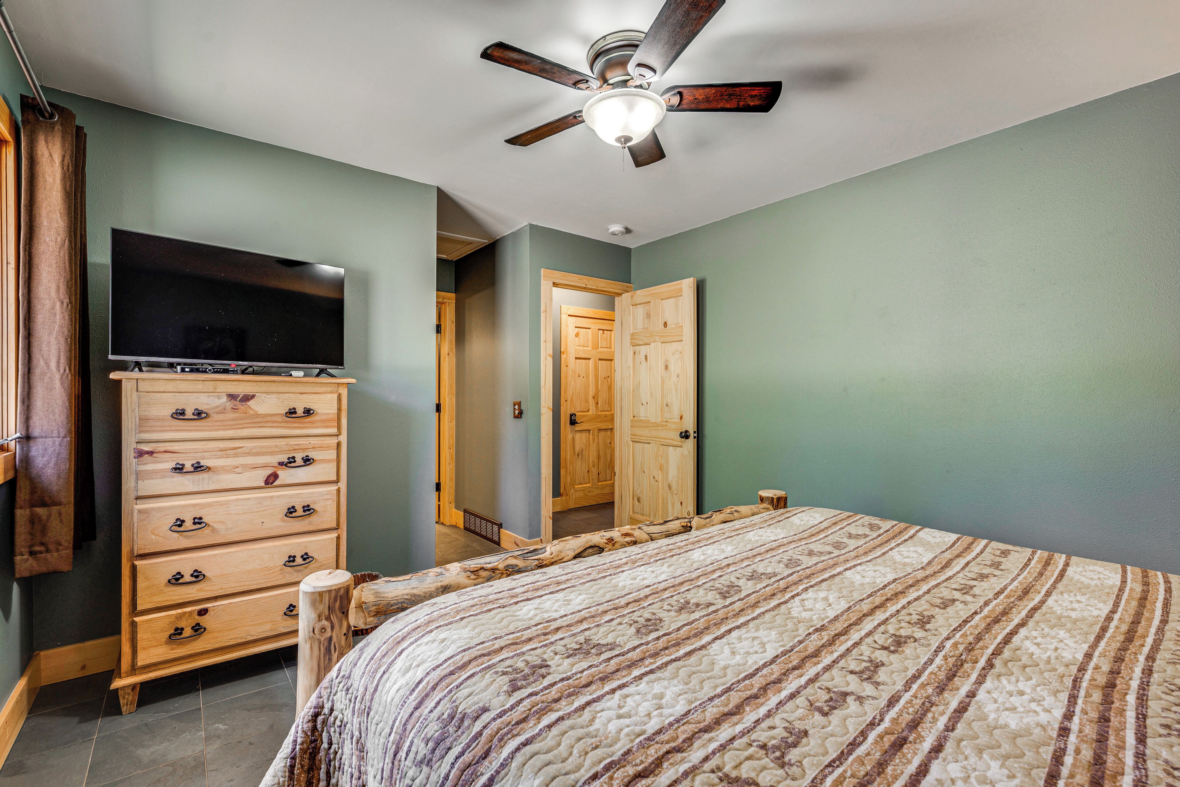 A bedroom with a bed, a dresser, a television, a ceiling fan, and a wooden door.