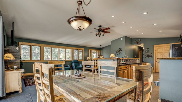 A spacious dining area with a large wooden table and chairs, a ceiling fan, and a view of the outdoors through large windows. - Estes Park Home w/ AC - 1/2 Mi to Lake & Downtown! (Estes Park)