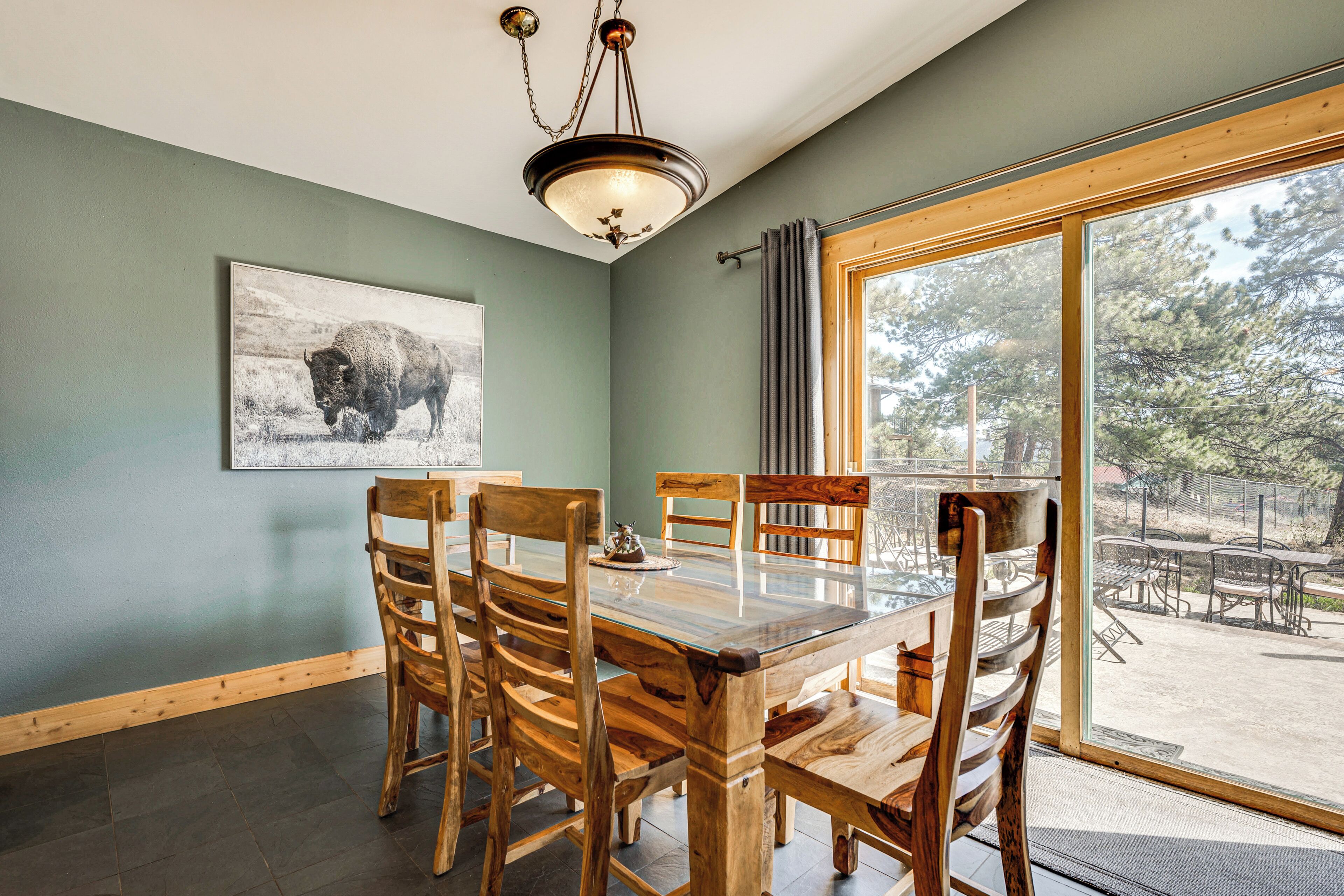 A dining area with a glass-top table, wooden chairs, a chandelier, a framed picture on the wall, and a large window with a view of the outdoors.