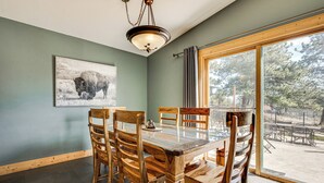 A dining area with a glass-top table, wooden chairs, a chandelier, a framed picture on the wall, and a large window with a view of the outdoors. - Estes Park Home w/ AC - 1/2 Mi to Lake & Downtown! (Estes Park)