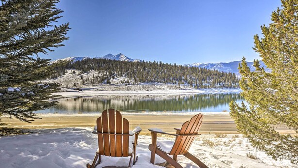 Two wooden Adirondack chairs facing a calm lake with snow-covered ground and a forested mountain range in the background. - Mtn/lake Views & 3 Mi to Skiing: Dillon Condo (Dillon)