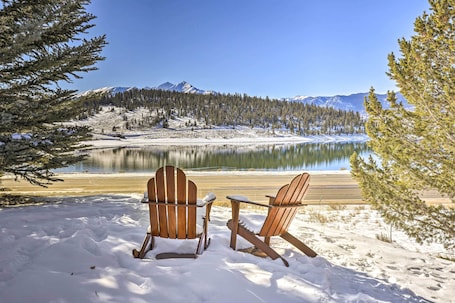Two wooden Adirondack chairs facing a snow-covered landscape with a lake and mountains in the background.