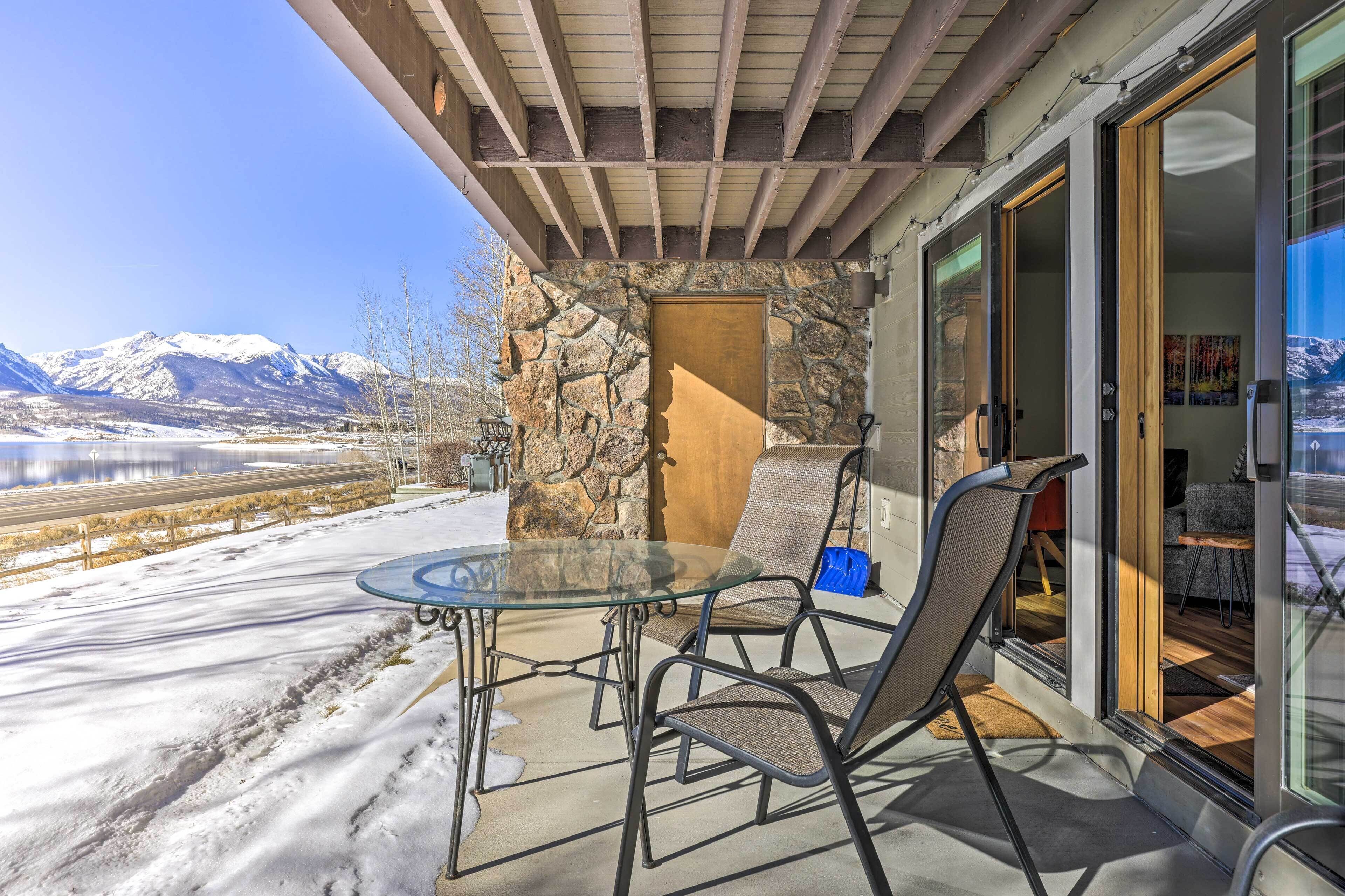 A patio with a glass table and chairs, a stone wall, and a view of mountains and a lake.