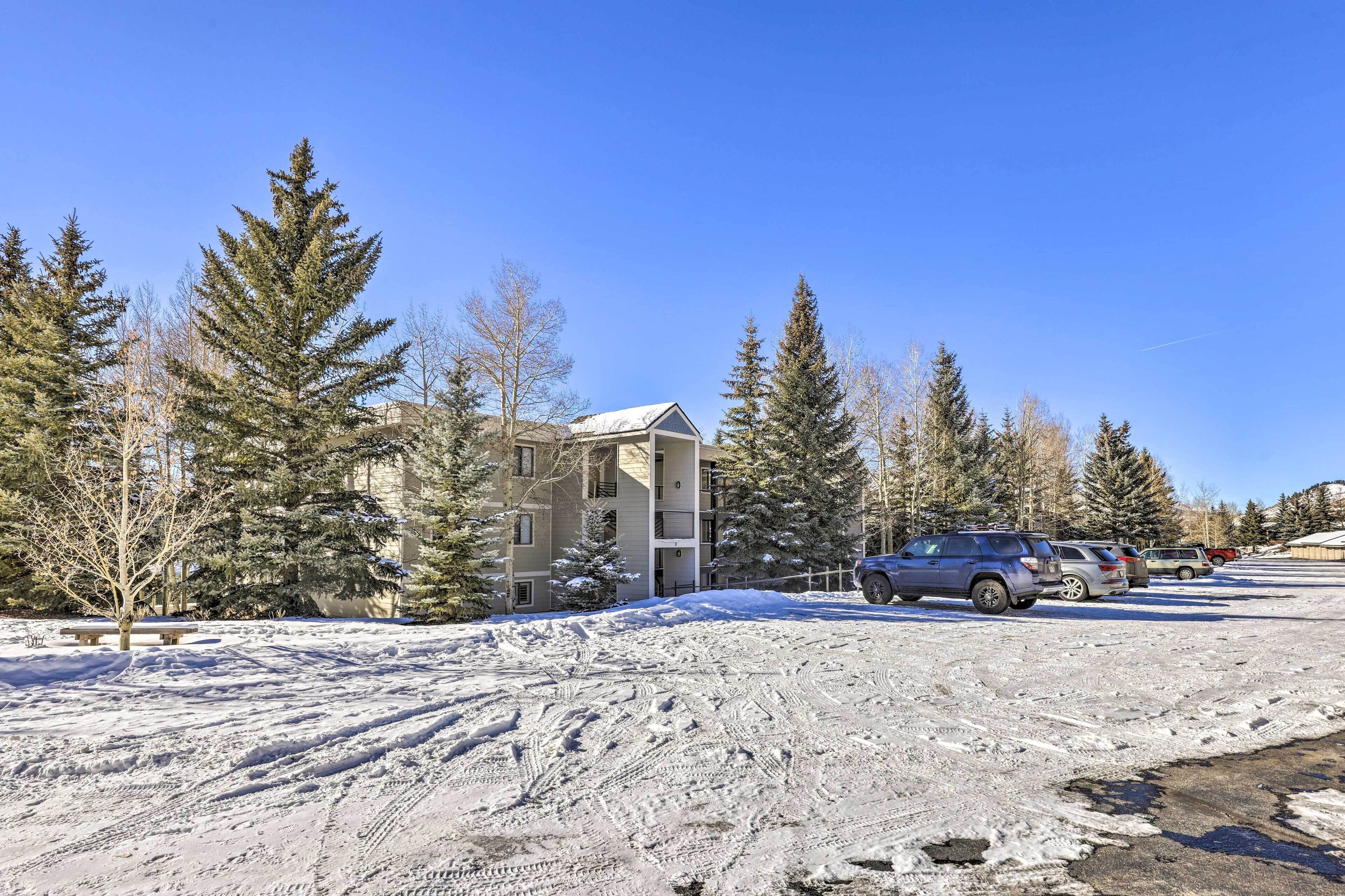 A parking lot with snow-covered ground, parked cars, and a building with a sloped roof in the background.