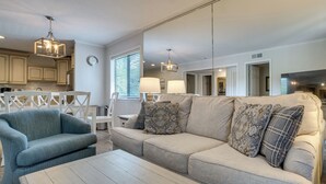 Ein modernes Wohnzimmer mit einem beigen Sofa, einem blauen Sessel und einem Holztisch. Zwei Pendelleuchten und ein großer Spiegel sind an der Wand angebracht.
