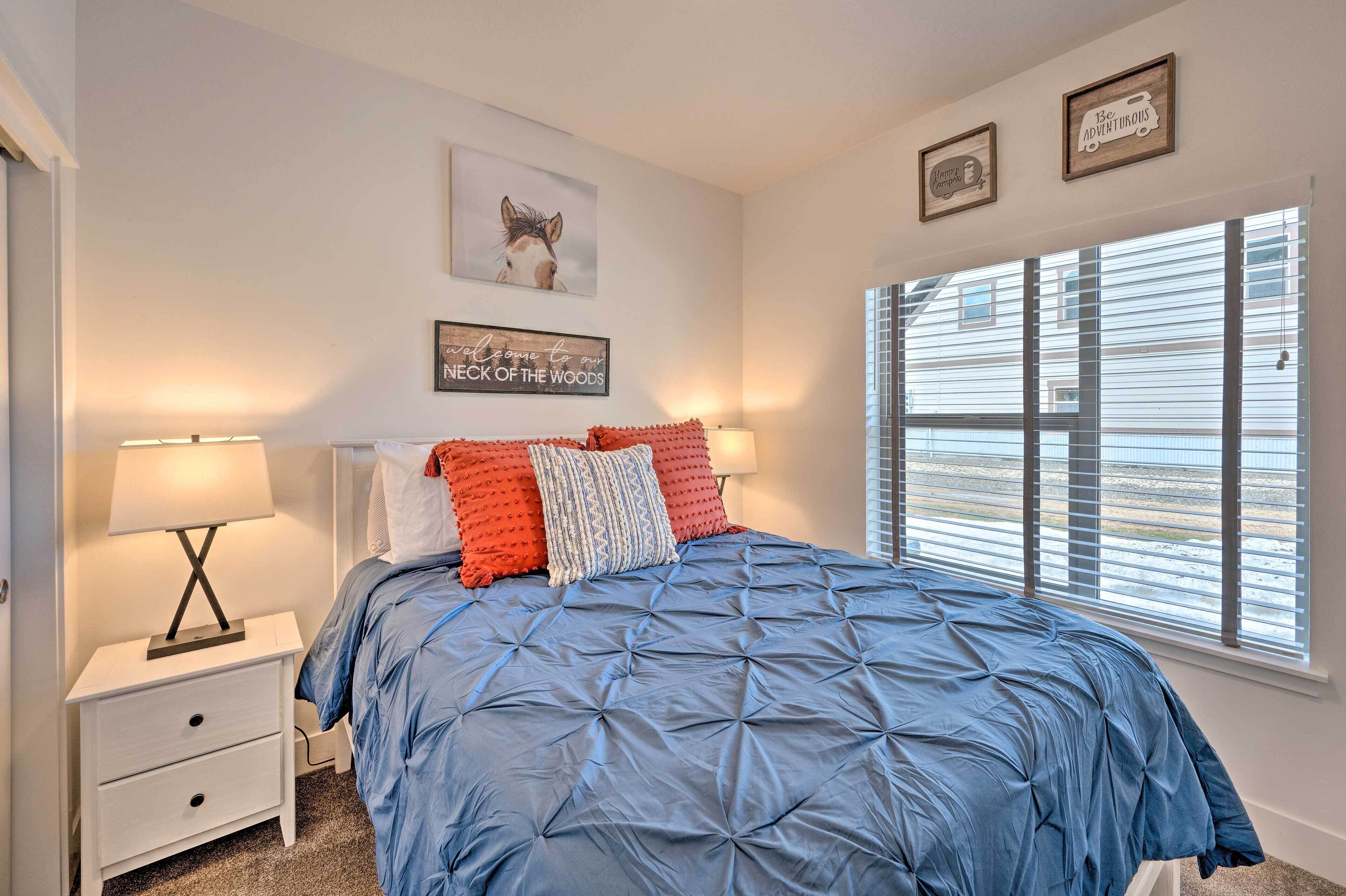 A bedroom with a blue quilted bedspread, a white nightstand with drawers, a lamp, and two framed pictures on the wall.
