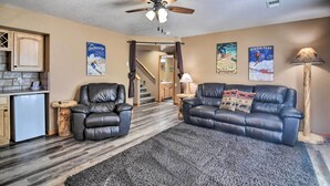 A living room with a ceiling fan, a fireplace, a sofa, a coffee table, and a lamp. There are posters on the wall and a staircase leading to another room. - Family-friendly Home w/ Hot Tub: 1 Mi to Dtwn (Estes Park)