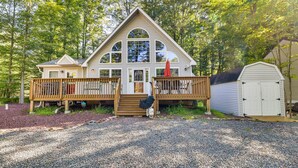 Une cabane à deux étages avec un porche à l’avant, une terrasse en bois et un abri de jardin.