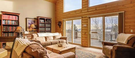 A spacious living room with a high ceiling, wooden walls, and large windows. There are two sofas, a coffee table, and a bookshelf filled with books.