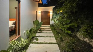 A modern house with a stone pathway leading to a wooden door, surrounded by lush greenery.