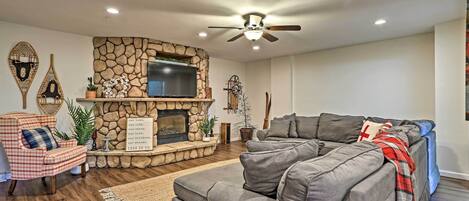 A living room with a stone fireplace, a television mounted above it, a grey sectional sofa, and a ceiling fan.