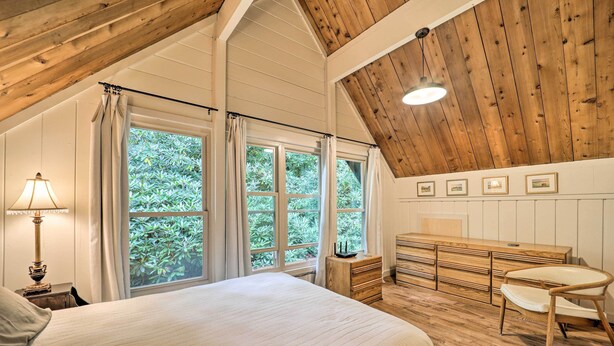 Une chambre à coucher avec un grand lit, une commode en bois, un bureau et une chaise. Il y a des fenêtres avec des rideaux, une lampe et des tableaux encadrés au mur.