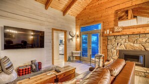 A log cabin interior with a stone fireplace, leather sofa, wooden coffee table, and a television mounted on the wall. - Pagosa Lakefront Home w/ Hot Tub, A/c, & Canoe! (Pagosa Springs)