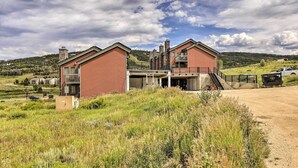 A modern two-story house with a balcony, surrounded by a grassy landscape and a dirt road. - Mtn View Granby Condo w/ In-unit Hot Tub! (Granby)