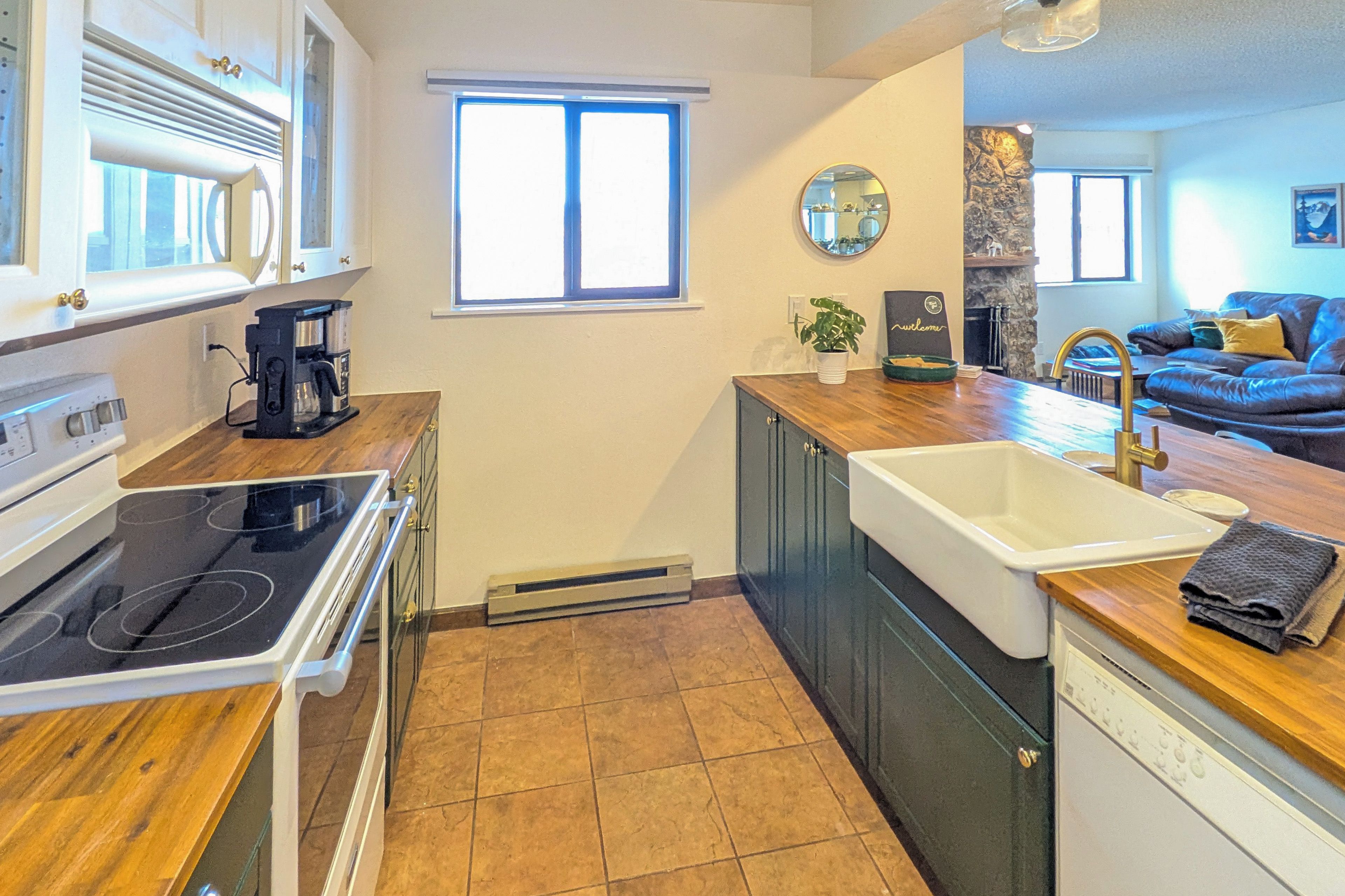 Cocina compacta con estufa, horno y lavamanos. Una ventana proporciona luz natural y vistas al área de estar con un sofá.